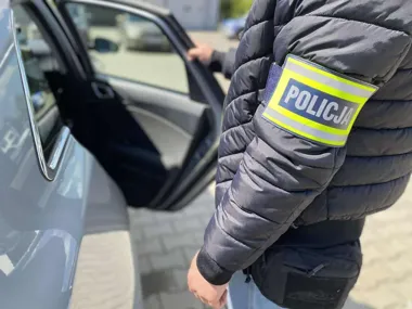Funkcjonariusz policji w czarnej kurtce z opaską „POLICJA” przy otwartych drzwiach samochodu.