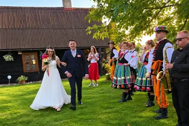 Para młoda przechodzi przez trawnik przed drewnianym domem, a obok stoją osoby w strojach ludowych oraz muzyk z saksofonem.